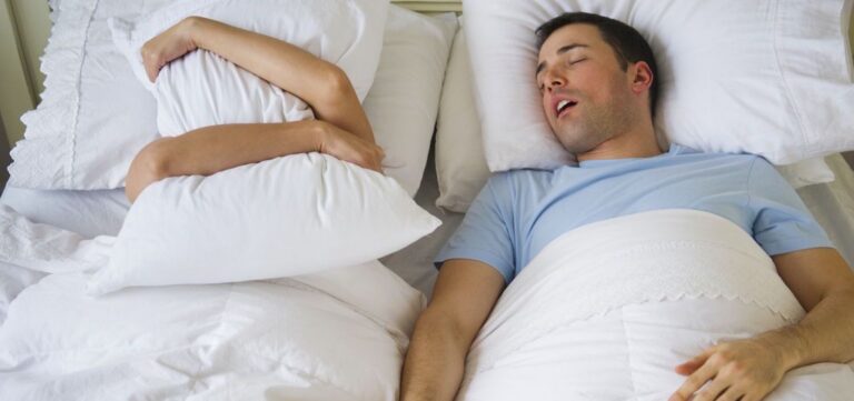 Bărbat care sforăie în pat, iar femeia de lângă el ține o pernă în fața capului, ilustrând cum sforăitul afectează viața de cuplu și soluții pentru această afecțiune