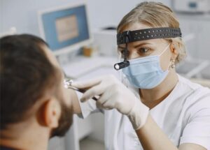 Consult ORL – medic specialist examinează urechea unui pacient în cabinet, folosind otoscop și echipament de protecție.